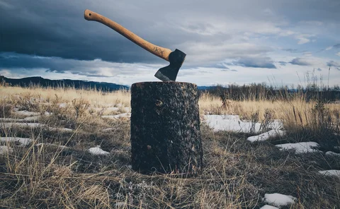 A wooden-handled sharp axe stuck in a wooden stump in the middle of a dry grass field under the cloudy sky